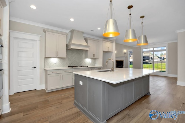 kitchen with premium range hood, sink, hanging light fixtures, a center island with sink, and decorative backsplash