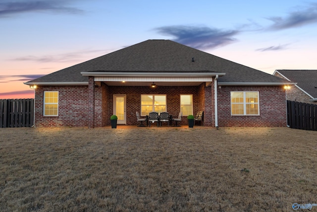 back of property at dusk featuring a lawn, roof with shingles, fence, a patio area, and brick siding