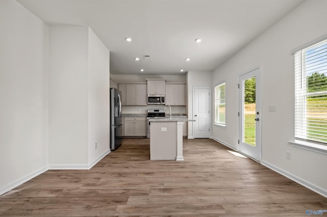 kitchen with appliances with stainless steel finishes, an island with sink, sink, light stone countertops, and light wood-type flooring