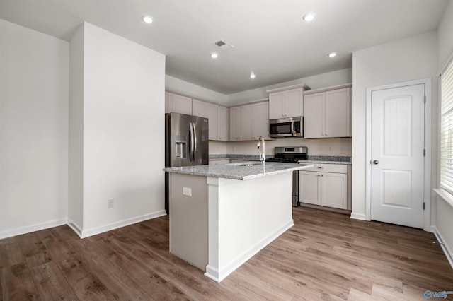 kitchen featuring sink, hardwood / wood-style floors, stainless steel appliances, light stone countertops, and an island with sink