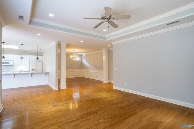 unfurnished living room with wood finished floors, visible vents, a tray ceiling, decorative columns, and ceiling fan with notable chandelier