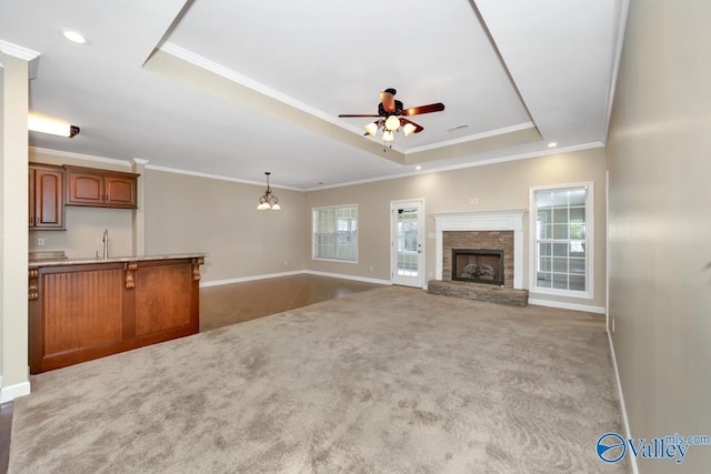 unfurnished living room featuring light carpet, ceiling fan with notable chandelier, a stone fireplace, crown molding, and a tray ceiling