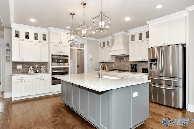 kitchen featuring appliances with stainless steel finishes, white cabinetry, an island with sink, custom exhaust hood, and light stone counters