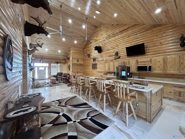 interior space featuring light brown cabinetry, wood walls, high vaulted ceiling, a center island with sink, and appliances with stainless steel finishes