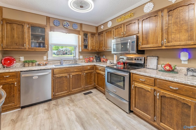 kitchen featuring crown molding, appliances with stainless steel finishes, a sink, brown cabinetry, and light wood-type flooring