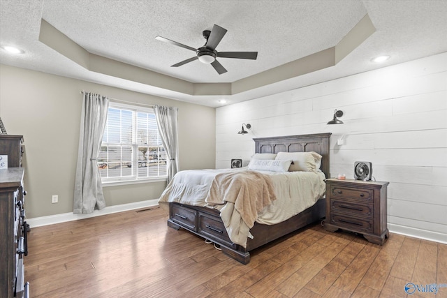 bedroom with a textured ceiling, a tray ceiling, ceiling fan, wooden walls, and wood-type flooring