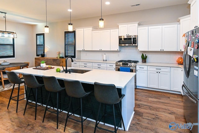 kitchen with sink, appliances with stainless steel finishes, a kitchen island with sink, white cabinets, and decorative light fixtures