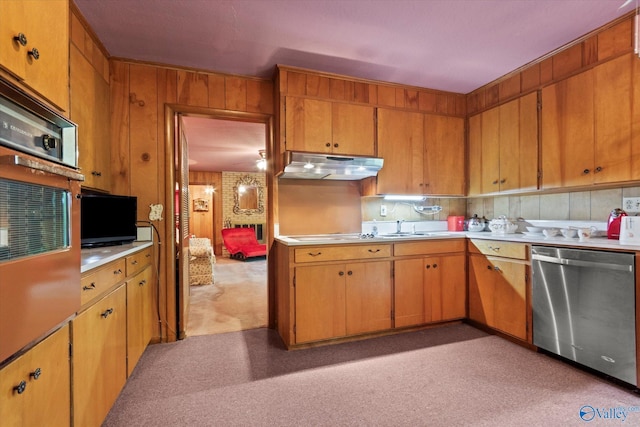 kitchen featuring brown cabinetry, under cabinet range hood, light countertops, dishwasher, and wooden walls