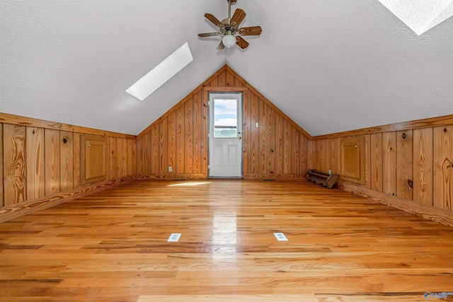 additional living space with wood walls, light hardwood / wood-style flooring, vaulted ceiling with skylight, and ceiling fan