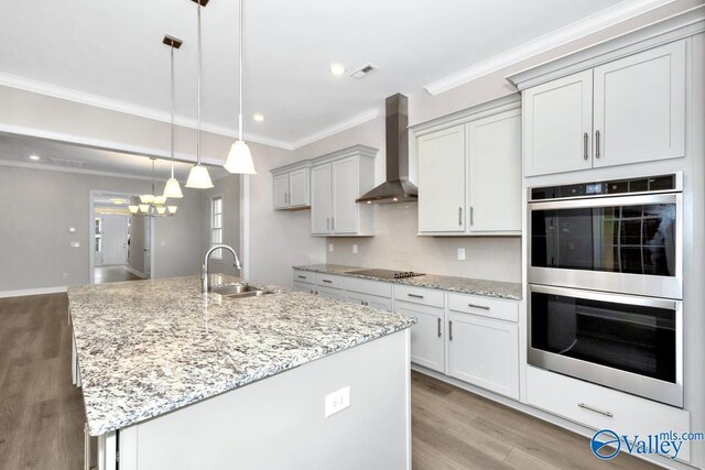 kitchen featuring double oven, decorative light fixtures, sink, wall chimney range hood, and a center island with sink