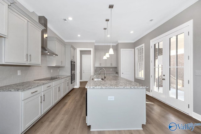 kitchen featuring sink, hanging light fixtures, a kitchen island with sink, light stone counters, and wall chimney range hood