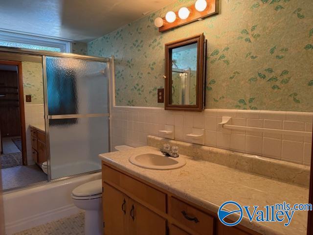 full bathroom featuring wallpapered walls, bath / shower combo with glass door, wainscoting, toilet, and vanity