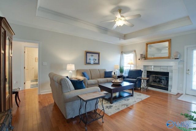 living room featuring ornamental molding, wood-type flooring, a raised ceiling, ceiling fan, and a tile fireplace