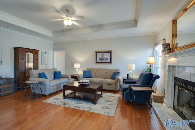 living room with a ceiling fan, a tray ceiling, a fireplace, ornamental molding, and light wood-type flooring