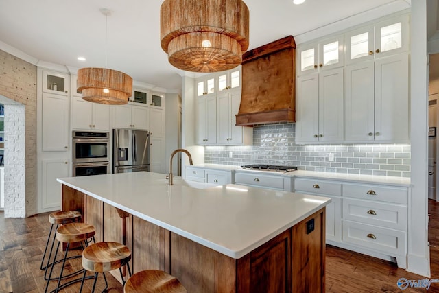 kitchen with light countertops, appliances with stainless steel finishes, glass insert cabinets, a kitchen island with sink, and white cabinetry