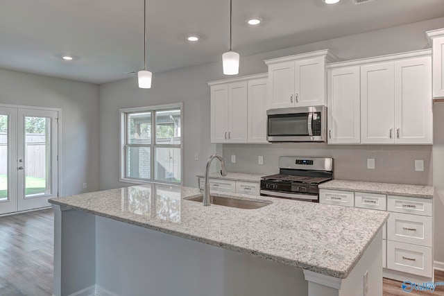 kitchen with appliances with stainless steel finishes, a kitchen island with sink, sink, white cabinets, and hanging light fixtures