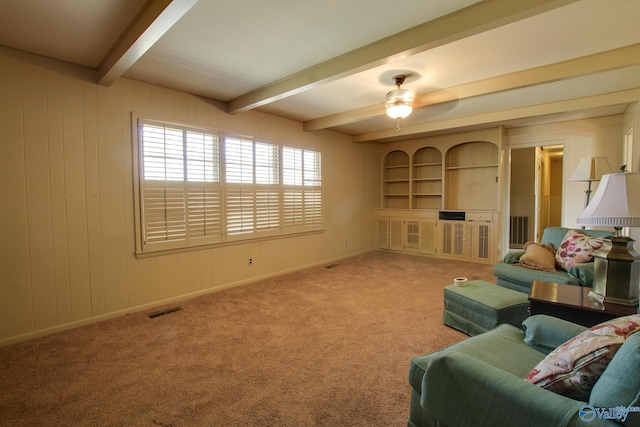 living room featuring built in shelves, wood walls, beamed ceiling, ceiling fan, and carpet