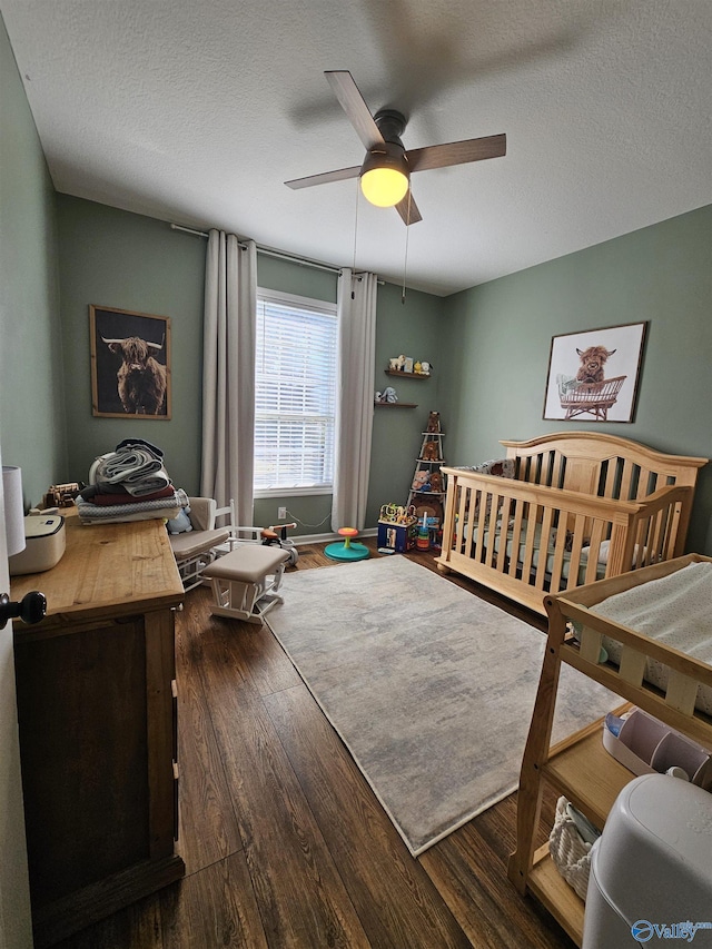 bedroom with dark wood-style floors, a textured ceiling, a nursery area, and a ceiling fan