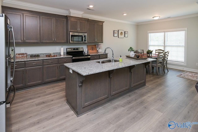 kitchen featuring appliances with stainless steel finishes, sink, light stone counters, light wood-type flooring, and an island with sink