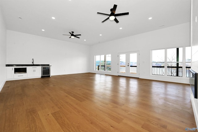 unfurnished living room with sink, ceiling fan, beverage cooler, french doors, and light wood-type flooring