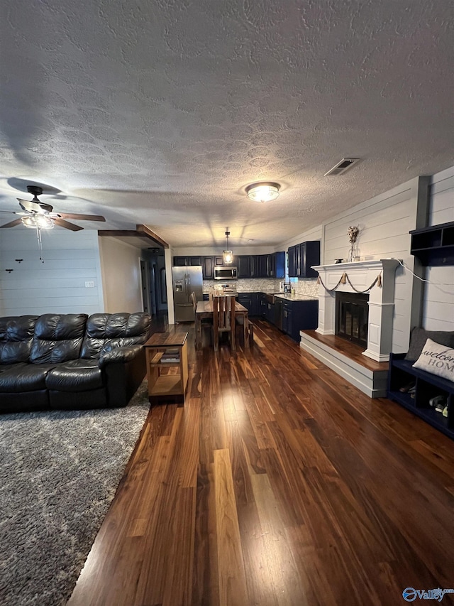 living room featuring a textured ceiling, ceiling fan, dark wood-type flooring, visible vents, and a glass covered fireplace