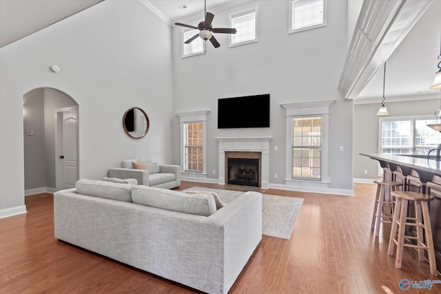 living area featuring arched walkways, a fireplace with flush hearth, ceiling fan, crown molding, and light wood-type flooring