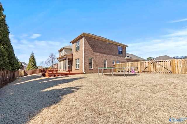 rear view of house with a deck, a fenced backyard, brick siding, a lawn, and a trampoline