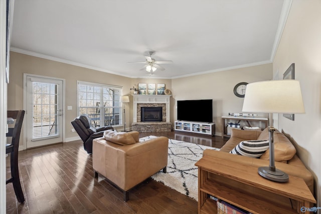 living area featuring a ceiling fan, dark wood finished floors, a stone fireplace, crown molding, and baseboards