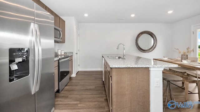 kitchen featuring stainless steel appliances, a kitchen island with sink, a sink, dark wood finished floors, and light stone counters