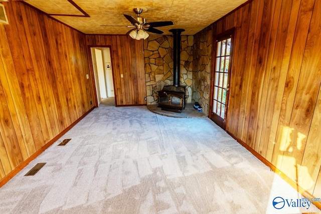 unfurnished living room featuring a wood stove, wooden walls, ceiling fan, and light carpet