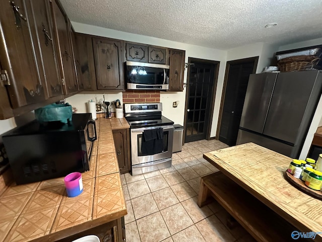 kitchen with a textured ceiling, dark brown cabinets, light tile patterned flooring, and stainless steel appliances