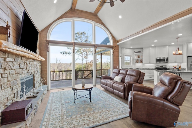 living room featuring a stone fireplace, ceiling fan, light hardwood / wood-style floors, high vaulted ceiling, and beam ceiling