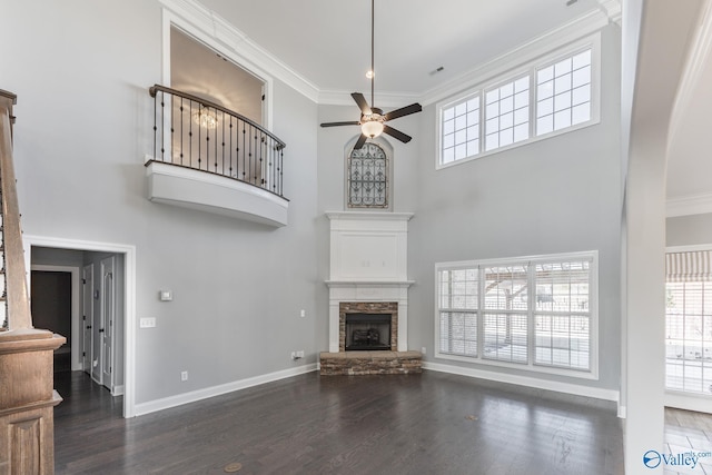 unfurnished living room featuring dark wood-type flooring, a high ceiling, a stone fireplace, crown molding, and ceiling fan