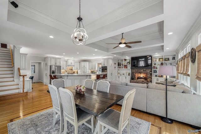 dining space with stairs, a stone fireplace, crown molding, a raised ceiling, and light wood-type flooring