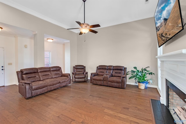 living room featuring a fireplace with flush hearth, wood finished floors, a ceiling fan, baseboards, and ornamental molding