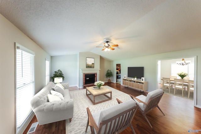 living room with a brick fireplace, wood-type flooring, ceiling fan with notable chandelier, vaulted ceiling, and a textured ceiling