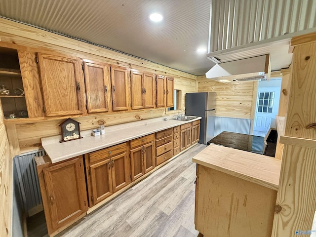 kitchen with brown cabinets, light wood-type flooring, wood walls, a sink, and freestanding refrigerator