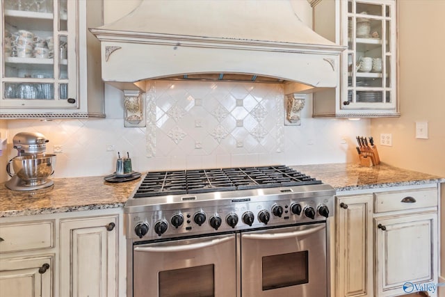 kitchen with backsplash, custom range hood, range with two ovens, light stone counters, and cream cabinets