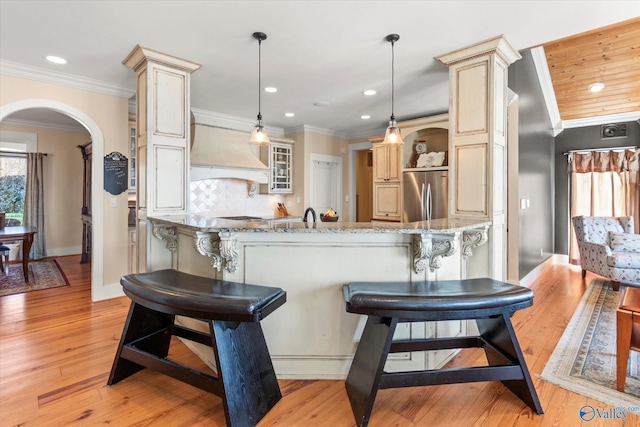 kitchen featuring light stone countertops, decorative backsplash, custom range hood, crown molding, and stainless steel fridge