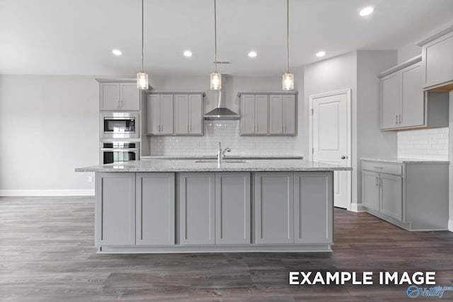 kitchen featuring appliances with stainless steel finishes, wall chimney range hood, a center island with sink, gray cabinets, and hanging light fixtures