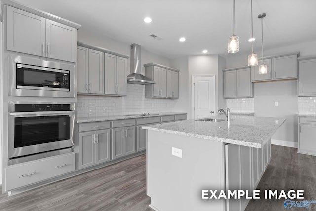 kitchen with sink, wall chimney exhaust hood, an island with sink, gray cabinets, and appliances with stainless steel finishes