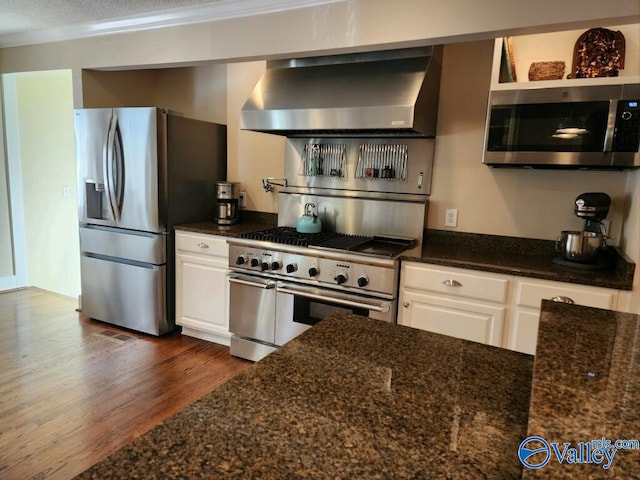 kitchen with wall chimney range hood, dark wood-type flooring, appliances with stainless steel finishes, dark stone countertops, and white cabinets