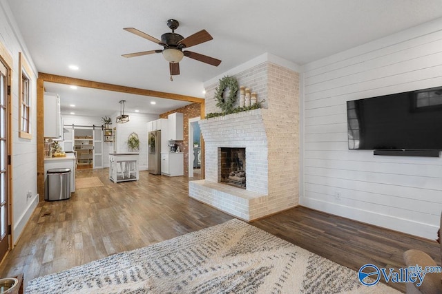 unfurnished living room featuring ceiling fan, a barn door, a brick fireplace, dark wood-type flooring, and brick wall