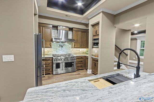 kitchen with a raised ceiling, decorative backsplash, ornamental molding, stainless steel appliances, and wall chimney range hood