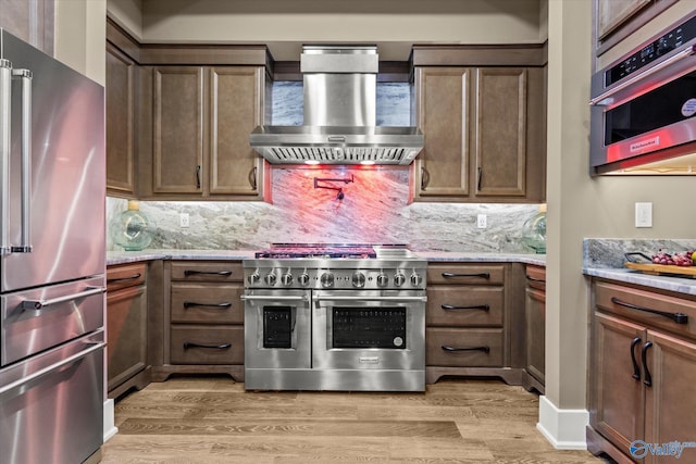 kitchen with high quality appliances, light stone countertops, wall chimney range hood, and light hardwood / wood-style floors