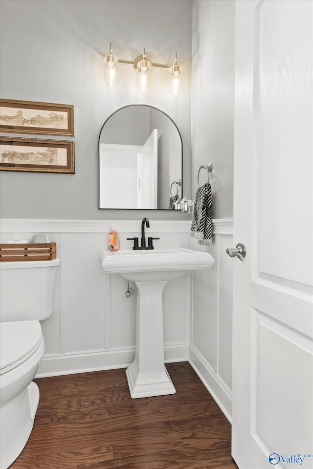 half bathroom featuring toilet, a decorative wall, wood finished floors, and wainscoting