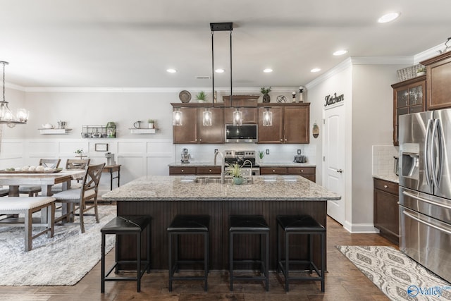 kitchen with light stone counters, dark brown cabinets, stainless steel appliances, a kitchen island with sink, and hanging light fixtures