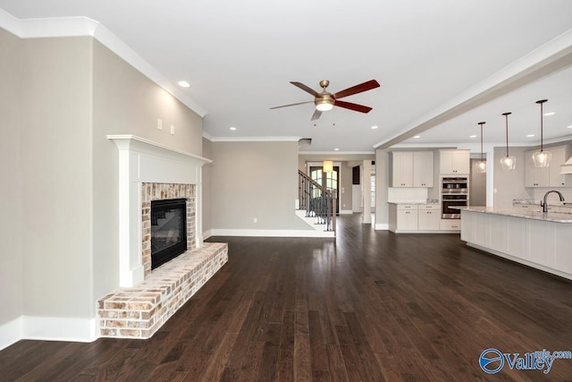 unfurnished living room with ornamental molding, dark wood-style floors, baseboards, a brick fireplace, and ceiling fan