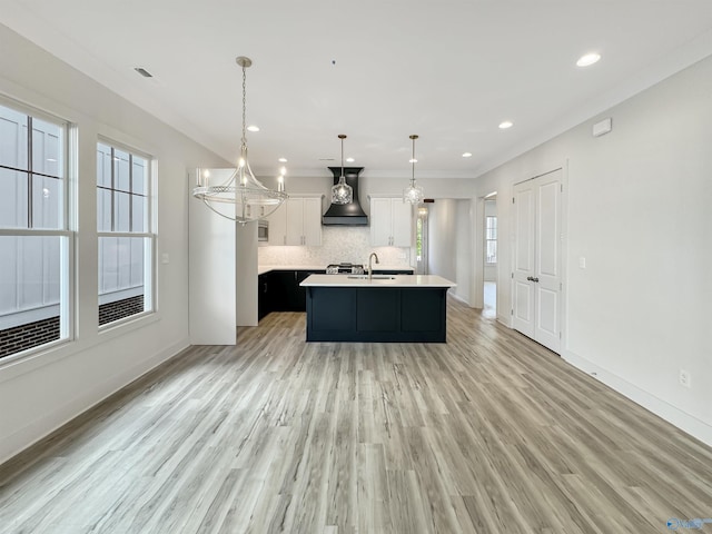 kitchen featuring pendant lighting, premium range hood, white cabinetry, backsplash, and an island with sink