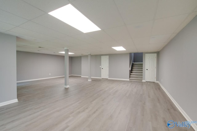 finished below grade area featuring stairs, light wood finished floors, a drop ceiling, and baseboards
